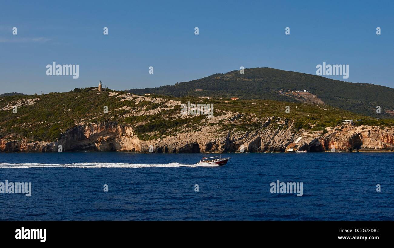 Isole IONIE, Zante, costa nord-orientale, Grotte Blu, Grotte Blu, luce del mattino, l'escursione in barca viaggia rapidamente da sinistra a destra, dietro le grotte, il faro, la costa rocciosa adornata, il mare blu scuro, cielo blu Foto Stock