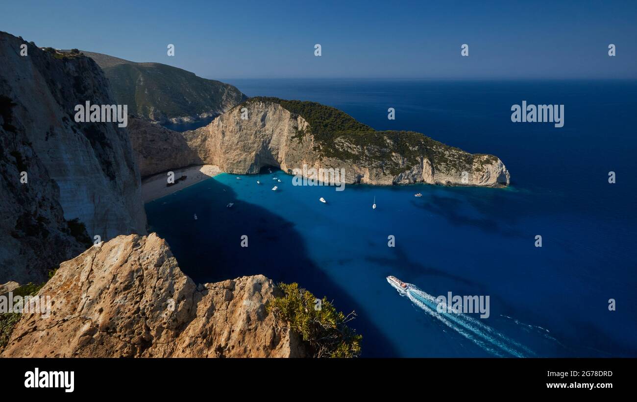 Zante, Paralia Navagio, Shipwreck Beach, vista grandangolare, vista dall'alto su tutta la baia e gran parte della spiaggia, diverse barche sono ancorate nella baia, grande barca da escursione si avvicina rapidamente dal basso a destra, massi illuminati dal sole in primo piano, acqua blu profondo, blu cielo Foto Stock