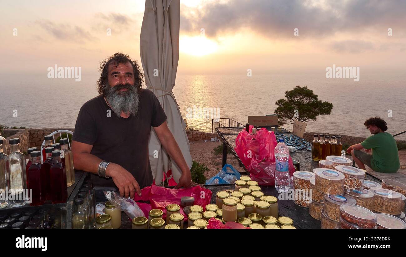 Zakynthos, Paralia Navagio, Shipwreck Beach, vendita stand per il miele ecc., greco uomo con barba, tramonto sullo sfondo, nuvole nel cielo Foto Stock