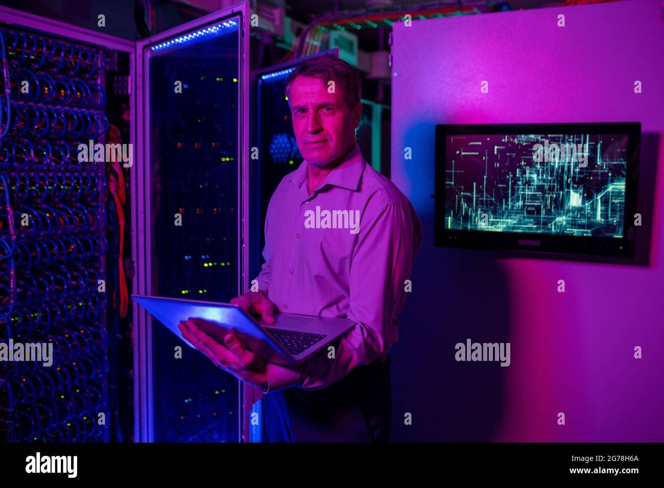 Ritratto di esperto IT maturo di contenuti in camicia con un notebook in un cabinet per server aperto nel data center Foto Stock