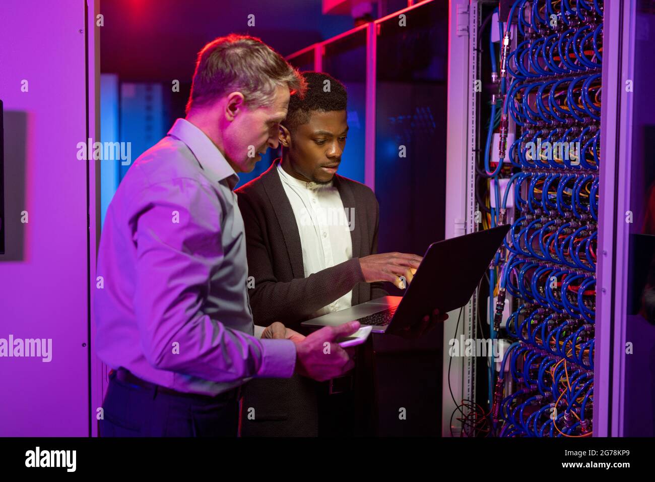 Team di specialisti di rete multietnici che si trova presso l'armadio server e utilizza un computer portatile durante il monitoraggio del traffico dei server Foto Stock