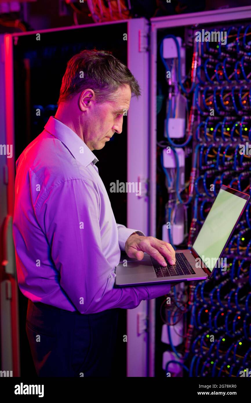 Tecnico IT caucasico concentrato e maturo che si trova a contatto con un cabinet per server in luce al neon e lavora con un notebook Foto Stock