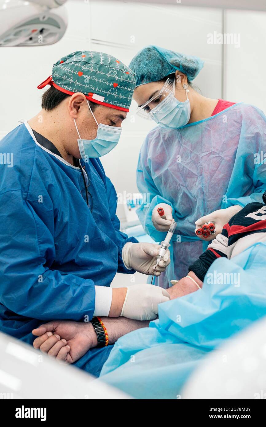 Smiley medico che lavora in clinica che preleva sangue a pazienti senza volto. Indossa una maschera facciale e indumenti protettivi. Foto Stock