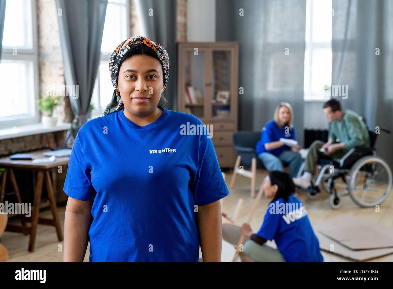 Volontaria femminile mista in t-shirt blu scuro in piedi contro i colleghi Foto Stock