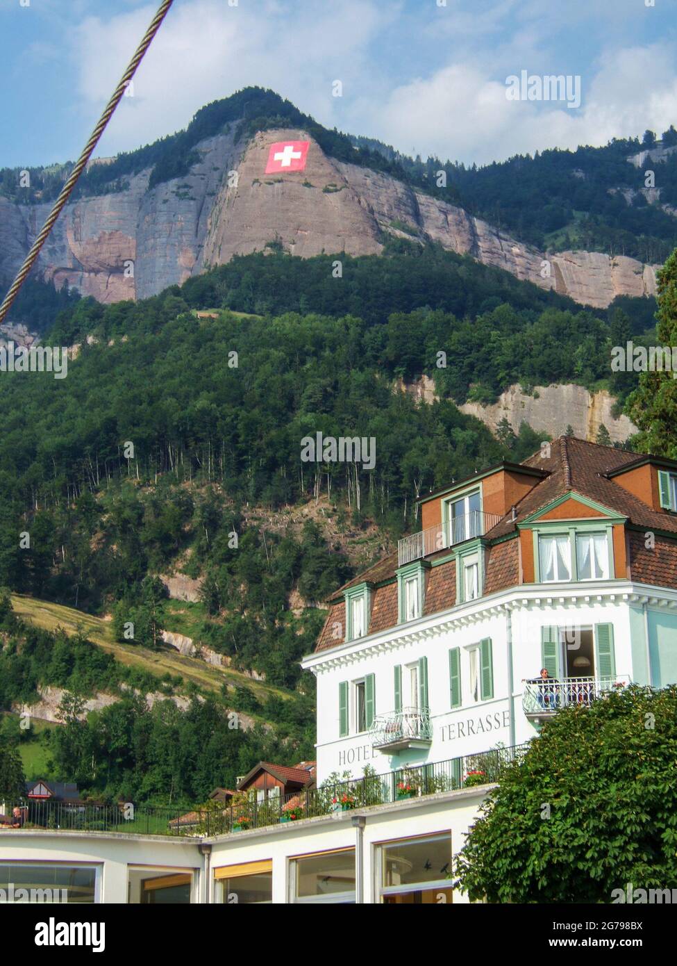 Lago di Lucerna, Svizzera: Agosto 10 2005: Un vecchio hotel bianco con persiane verdi di fronte ad una grande montagna con una parete rocciosa su cui una bandiera svizzera ca Foto Stock