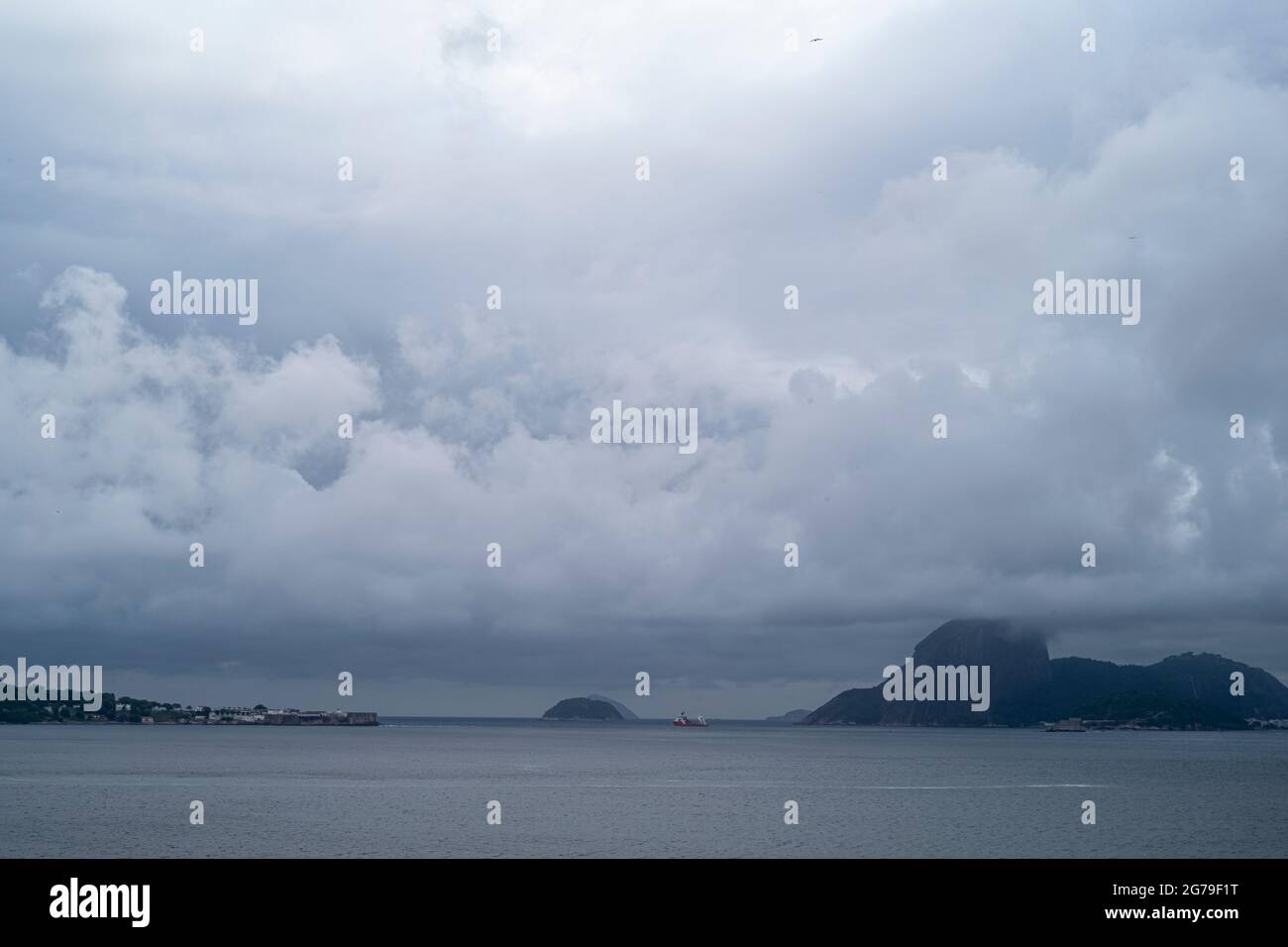 La vista da Ponte da Boa Viagem, Nitreói, Stato di Rio de Janeiro, Brasile. Isola verde Ilha da boa Viagem isola privata in baia collegata alla terra principale attraverso un ponte stretto e sabbia gialla sotto il paesaggio blu nuvoloso. Montagne in lontananza. Foto Stock