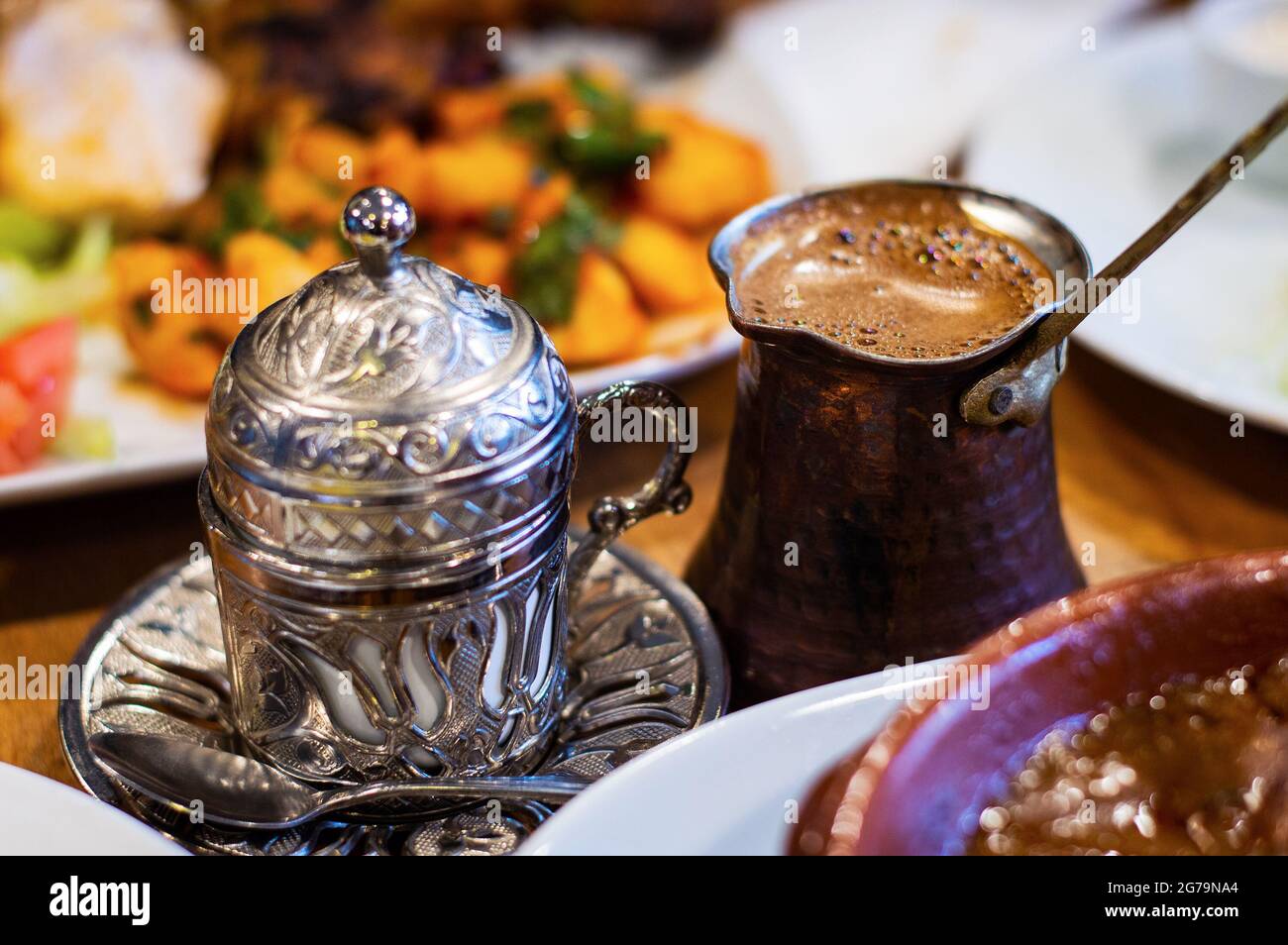 Caffè turco e tazza tradizionale in metallo goffrato sul tavolo. Cibo offuscato a Forground e background. Foto Stock
