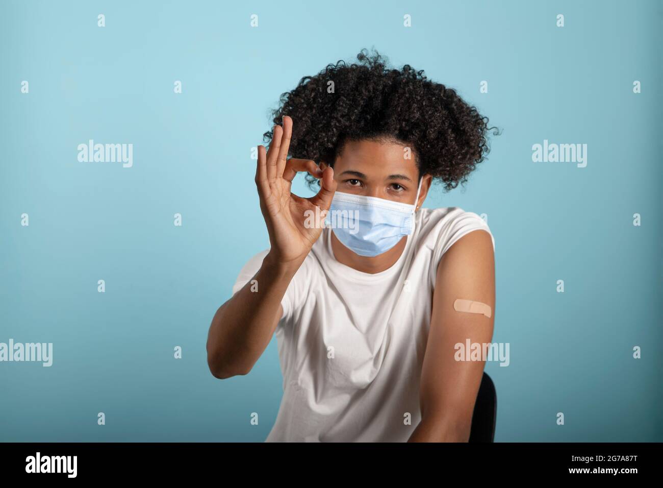 Giovane afrolatino che mostra braccio con coronavirus vaccino band-Aid con gesto ok e maschera protettiva su sfondo blu Foto Stock