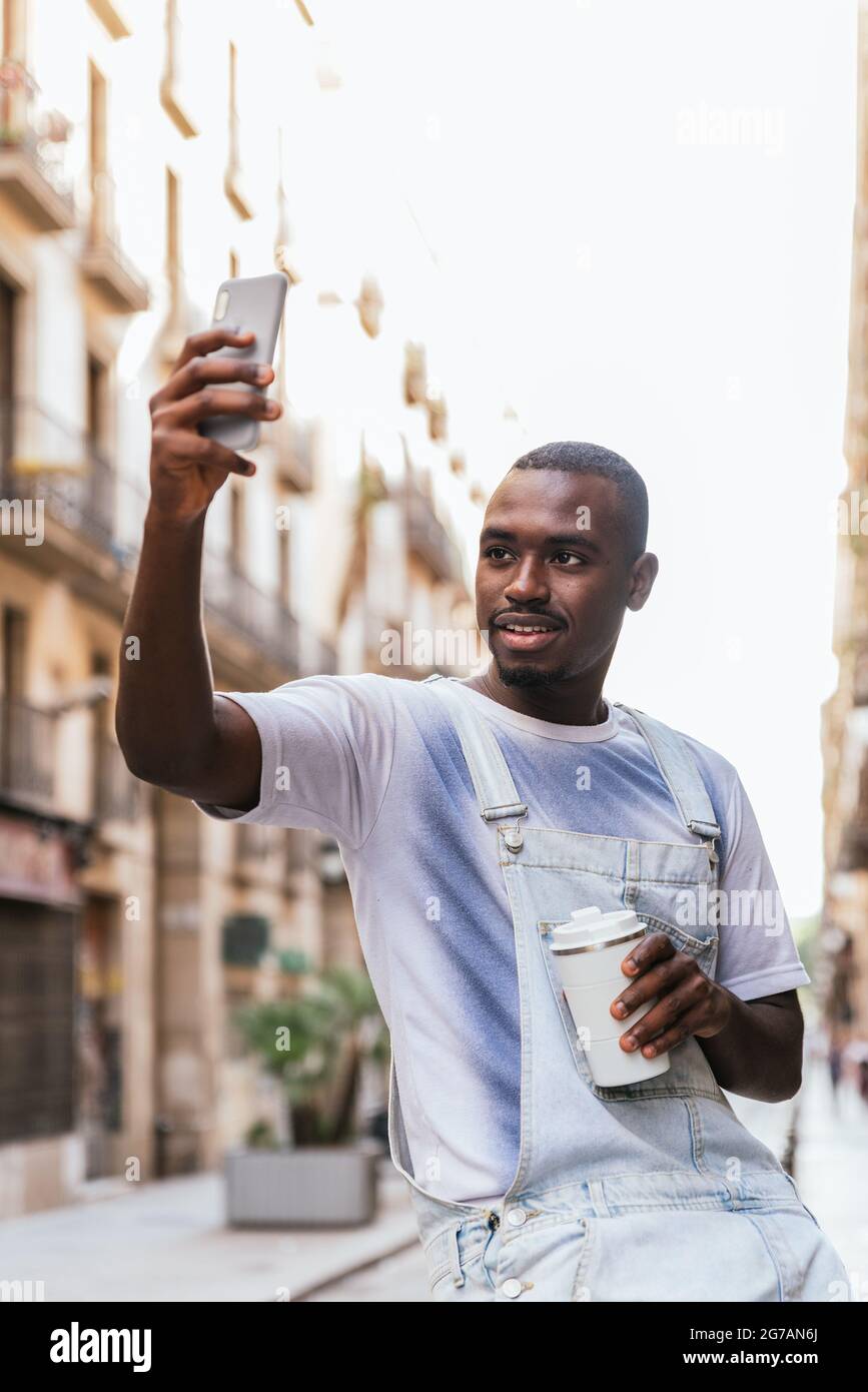 l'uomo attraente africano alzando il braccio con il suo telefono per prendere un selfie. Sorride alla macchina fotografica e tiene una tazza riutilizzabile di caffè Foto Stock
