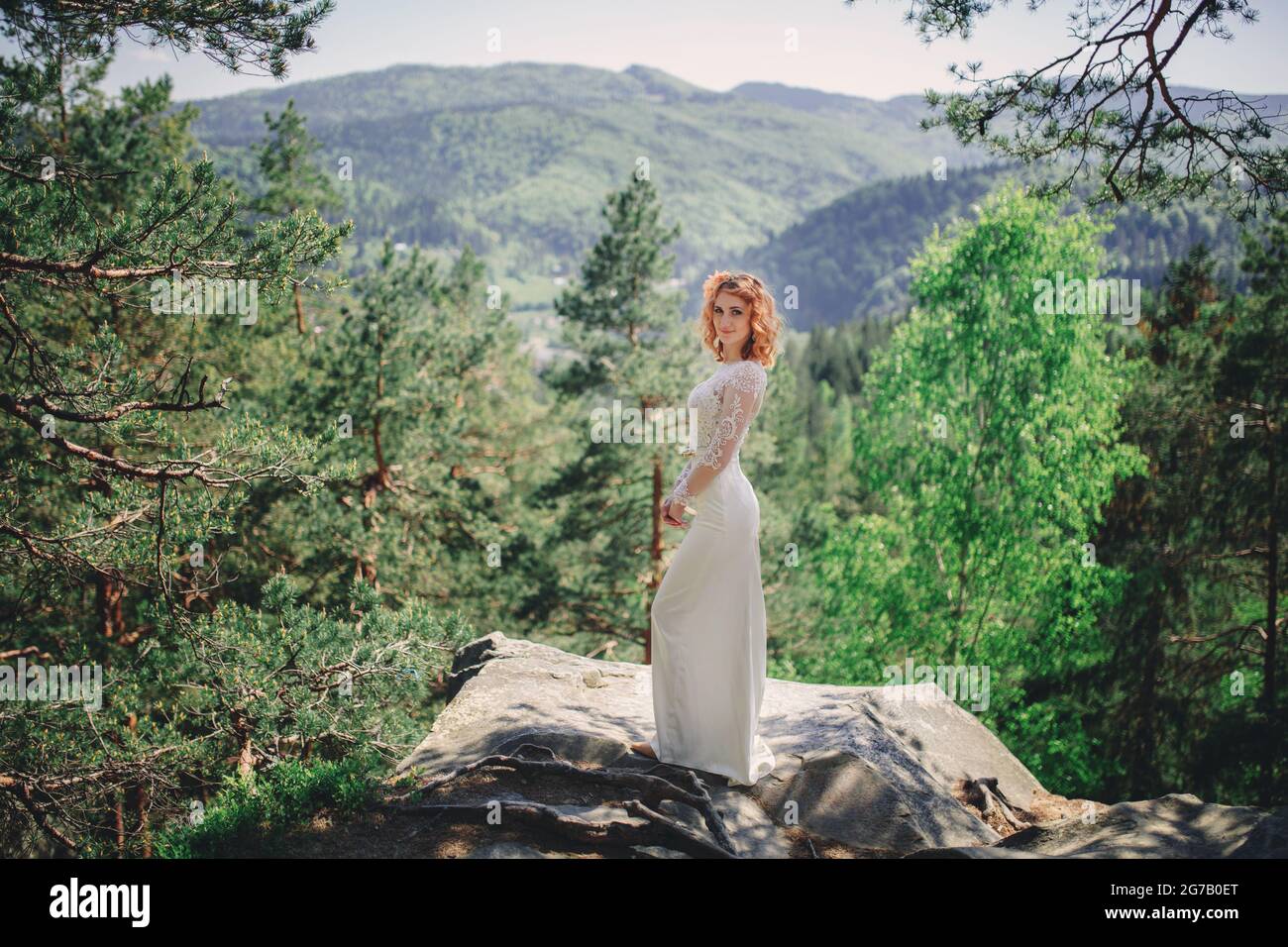 bella giovane sposa dai capelli rossi nella foresta con una corona floreale sulla testa. donna in lungo vestito bianco all'aperto il giorno d'estate. giorno di nozze. Foto Stock