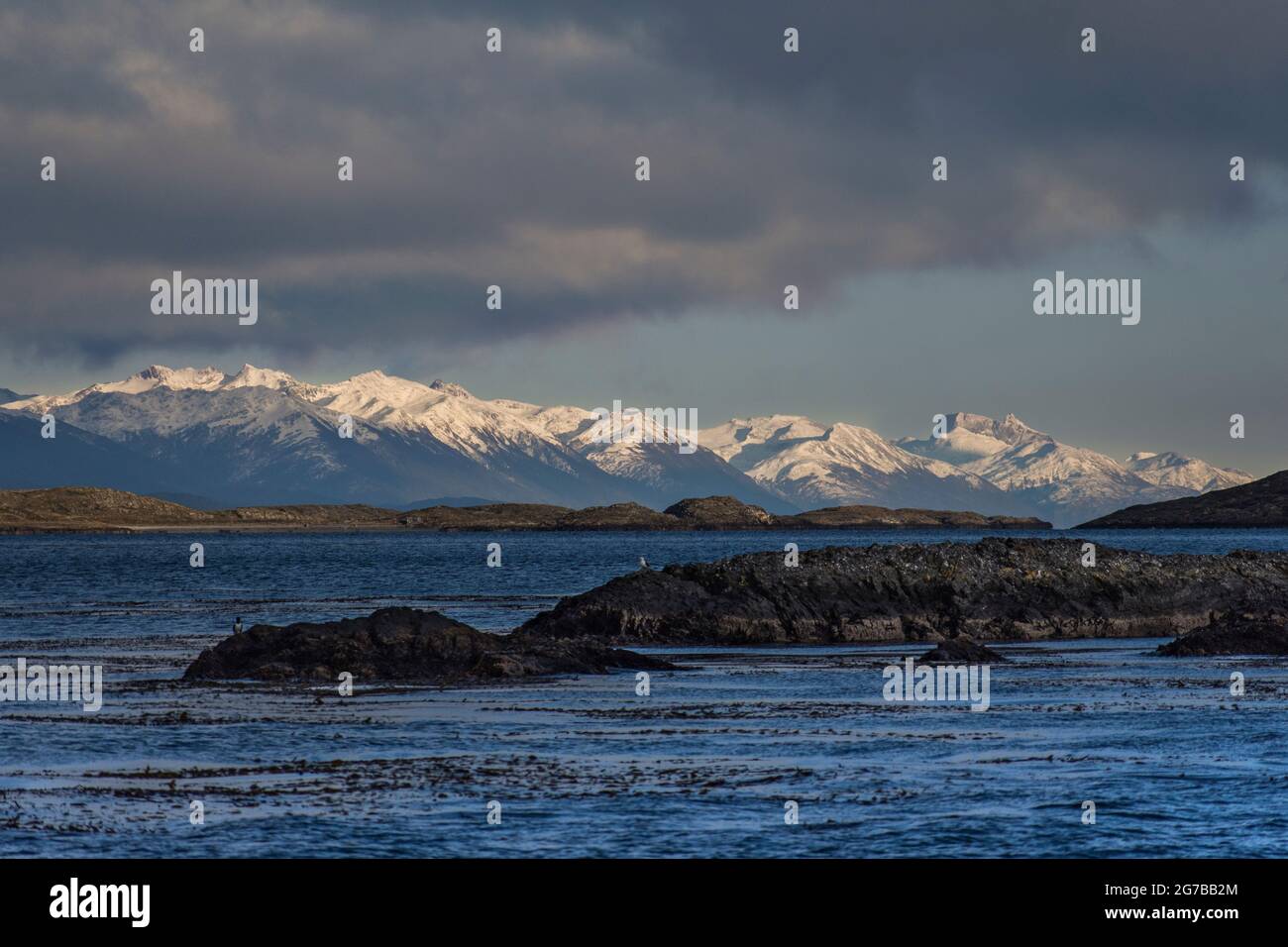 Scatto panoramico delle Ande ushuaiane innevate chiamate fine del mondo e di fronte una piccola isola desertica di roccia Foto Stock