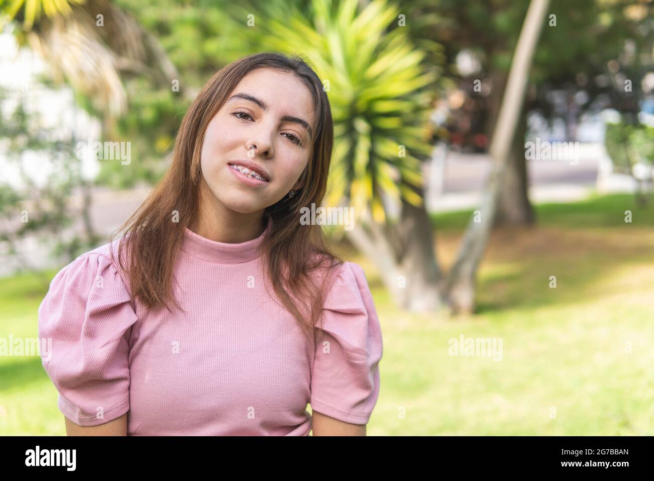Ritratto di una ragazza teenage latina con bretelle guardando la macchina fotografica all'aperto vestita in una blusa rosa in un parco Foto Stock
