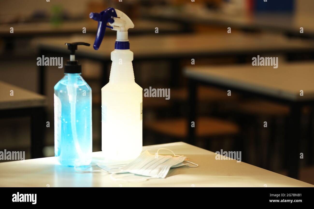 Un'aula scolastica vuota con scrivanie sullo sfondo. Una maschera per il viso, una bottiglia di disinfettante per le mani e uno spray per la pulizia si trovano in primo piano alla luce del sole Foto Stock