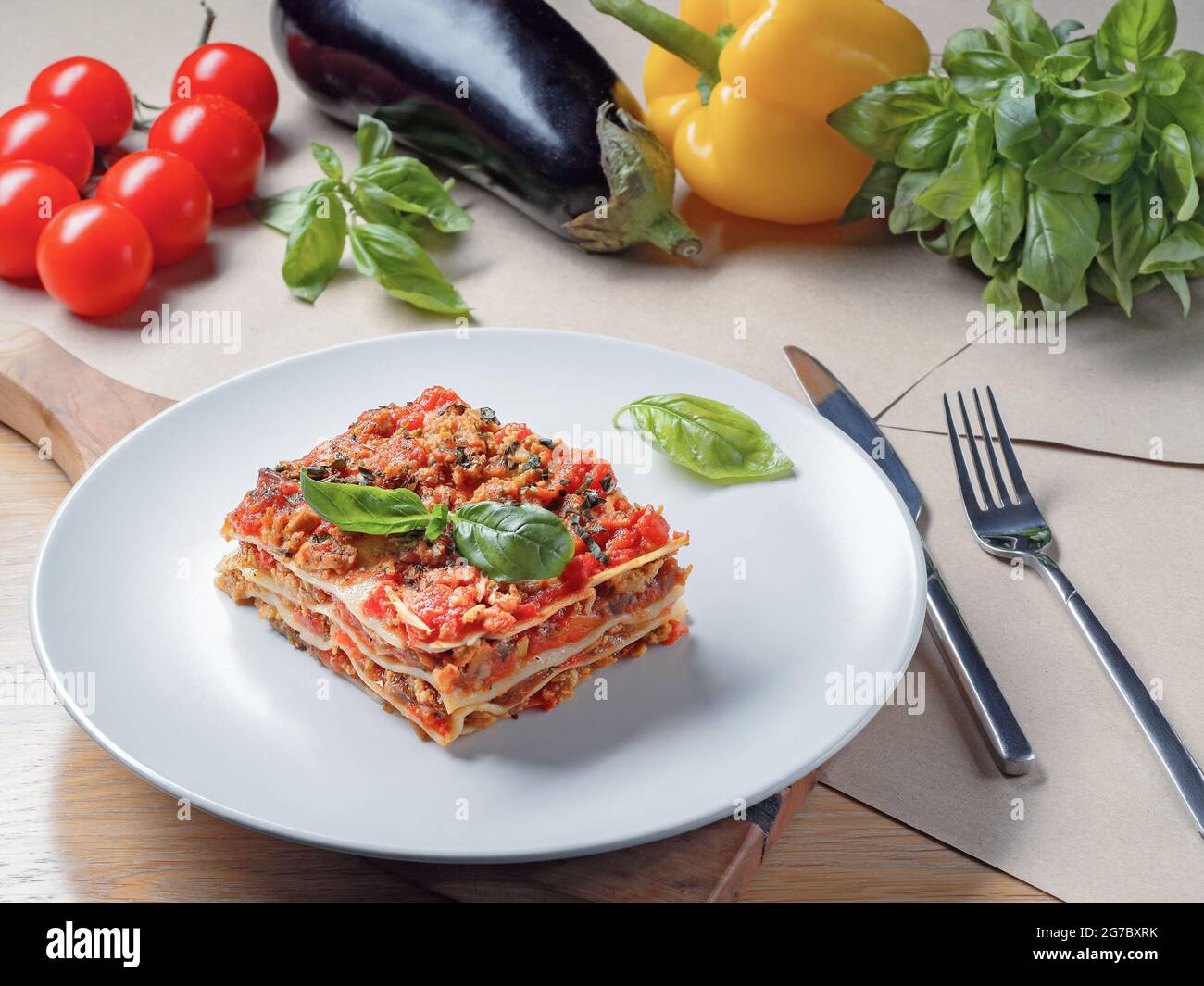 Pezzo di lasagna su piastra bianca. Basilico, pomodori, zucchine e pepe sullo sfondo. Menu di cucina Italiana. Foto Stock