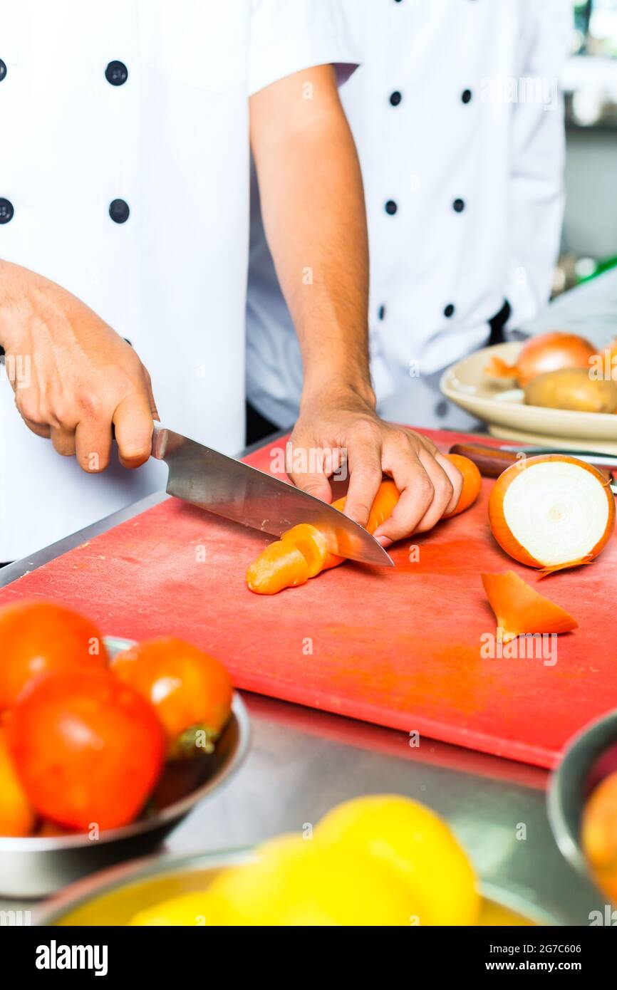Chef indonesiano asiatico insieme ad altri cuochi nel ristorante o nella cucina commerciale dell'hotel, affettando una carota Foto Stock