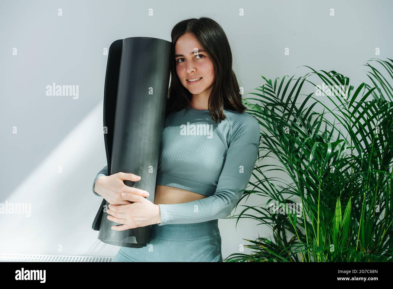 Giovane donna modesta con sorriso sottile che posa con un tappetino yoga, abbracciandolo. Foto Stock