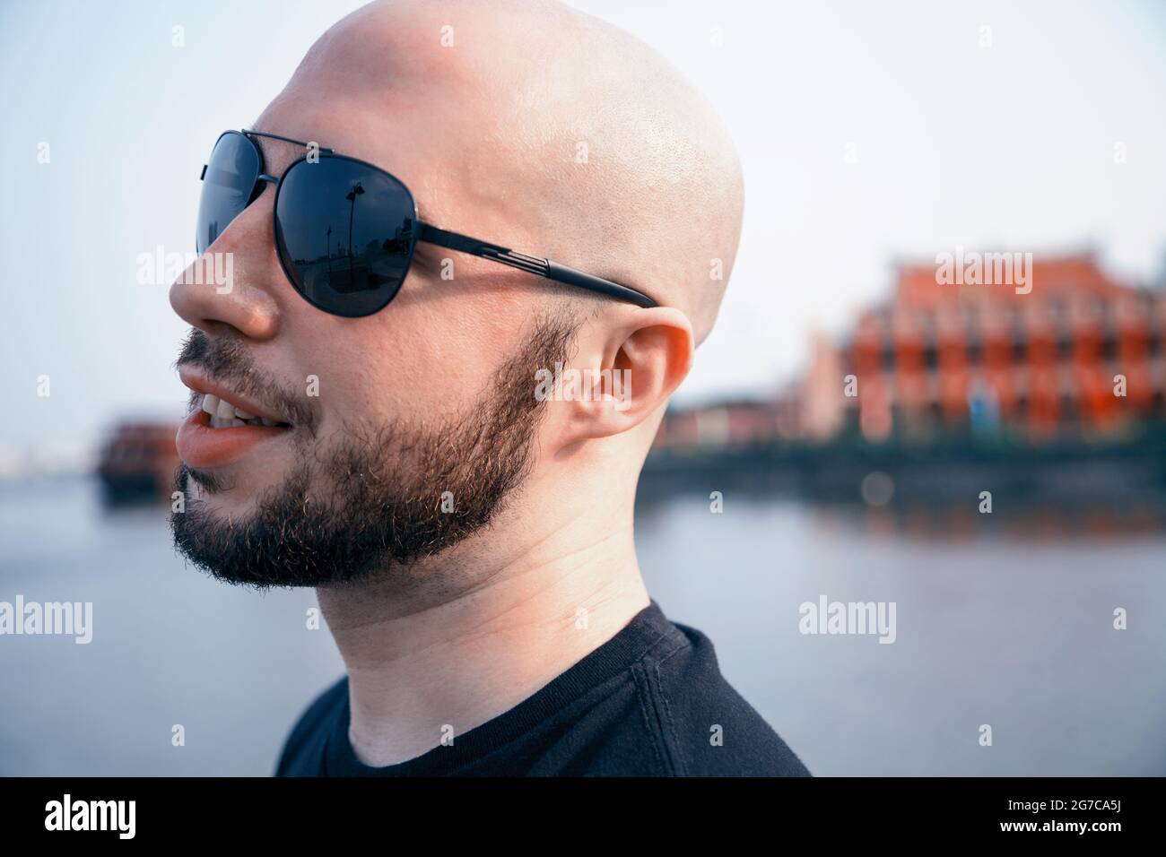 Felice giovane uomo in occhiali da sole che passeggiano lungo un fiume e  sognando. Ragazzo maschile calvo che sorride da vicino Foto stock - Alamy
