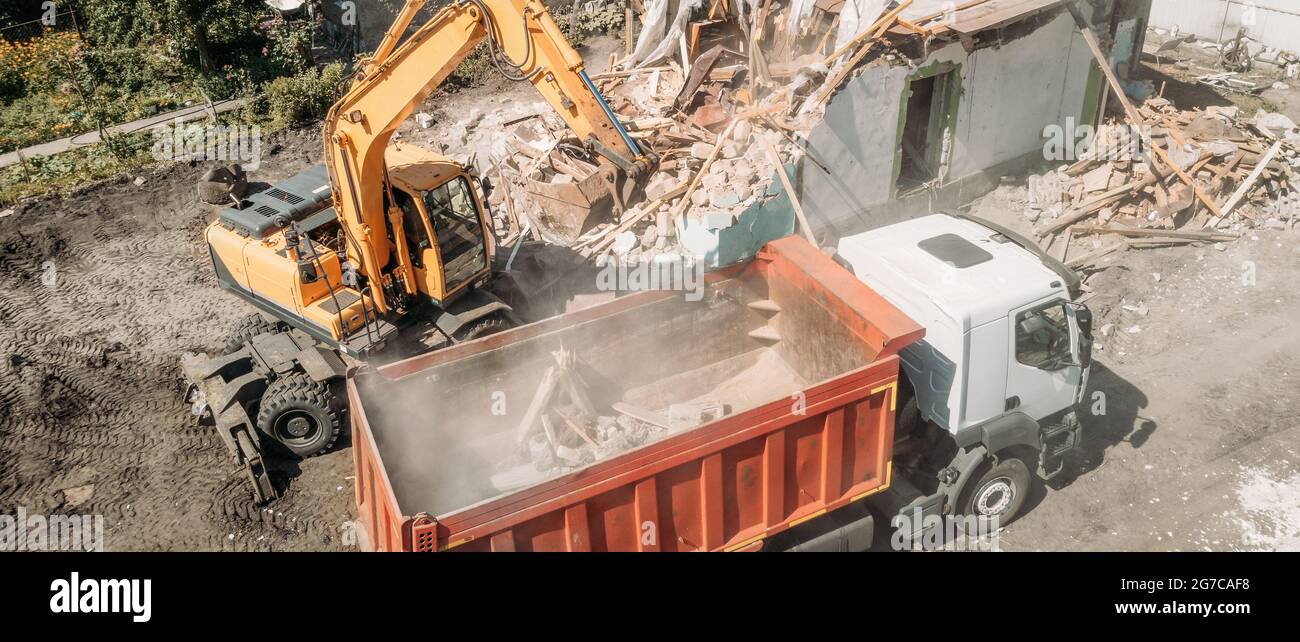 L'escavatore carica i detriti di costruzione del vecchio edificio dopo la distruzione nel dumper, vista aerea dal drone Foto Stock