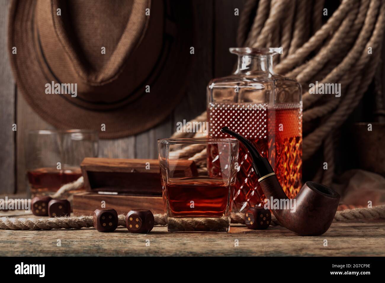 Whisky, pipe e dadi su un vecchio tavolo di legno. Sullo sfondo c'è una corda di canapa e un cappello da uomo. Foto Stock