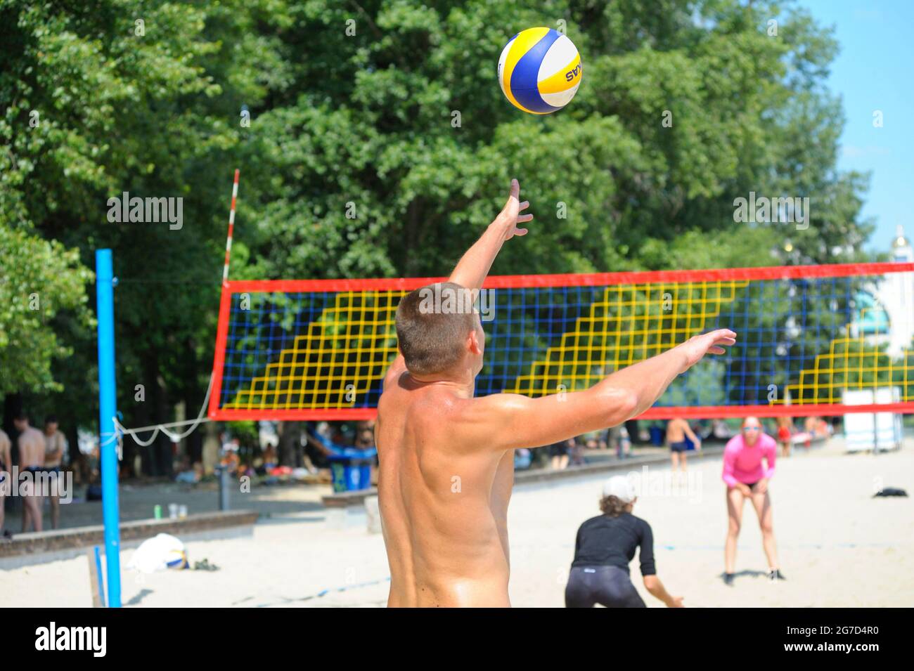 Beach volley. Giocatore di pallavolo maschile che serve la palla davanti alla rete, vista posteriore, giocatori sfocati. Foto Stock