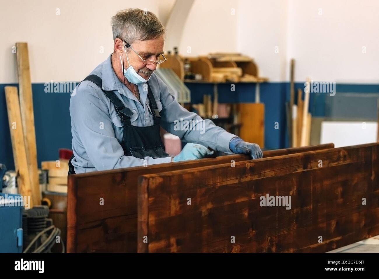 Un uomo anziano dipinge tavole di legno con vernice scura in un laboratorio di Falegnameria. Un pensionato in una maschera medica guadagna un lavoro part-time come un cabinetmaker. Lavoro fisico Foto Stock