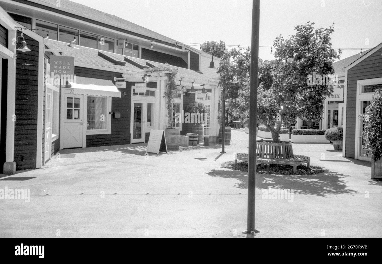 Fotografia in bianco e nero di storefronts, alberi e una panca rotonda in una piccola area commerciale a Larkspur Landing, Marin Country Mart, Larkspur, California, agosto 15, 2020. () Foto Stock