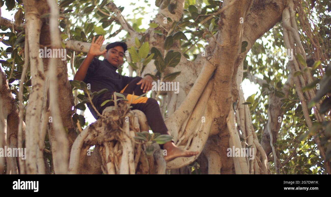 Un giovane ragazzo dell'Asia del Sud seduto su un grande albero che sventola a piedi nudi Foto Stock