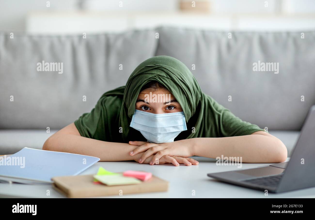 Ragazza teen musulmana in maschera e hijab che giace sul tavolo accanto al laptop e materiali di studio, sentendosi annoiato alla lezione online Foto Stock