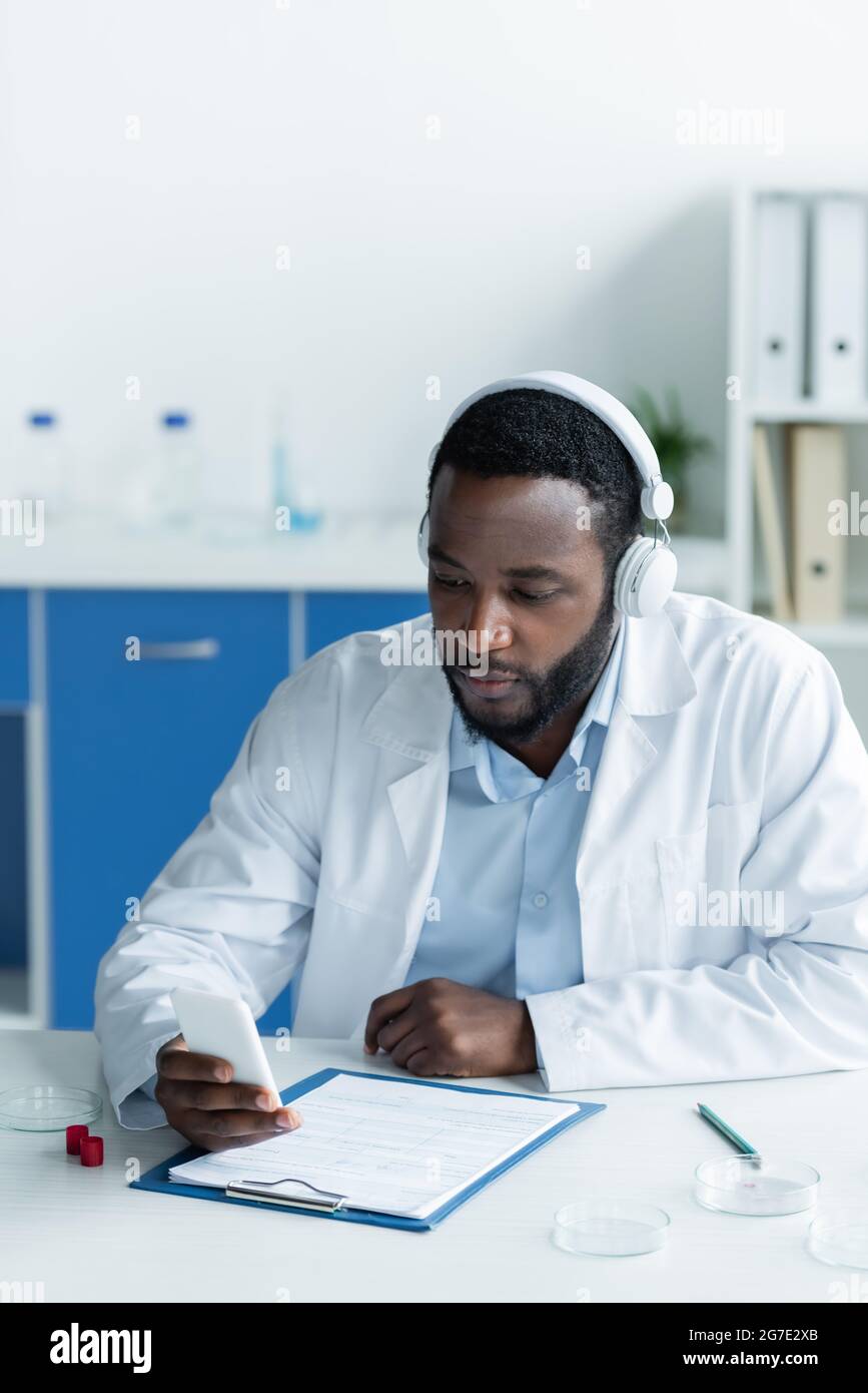 Scienziato afro-americano in cuffie con smartphone vicino a piatti Petri e appunti Foto Stock