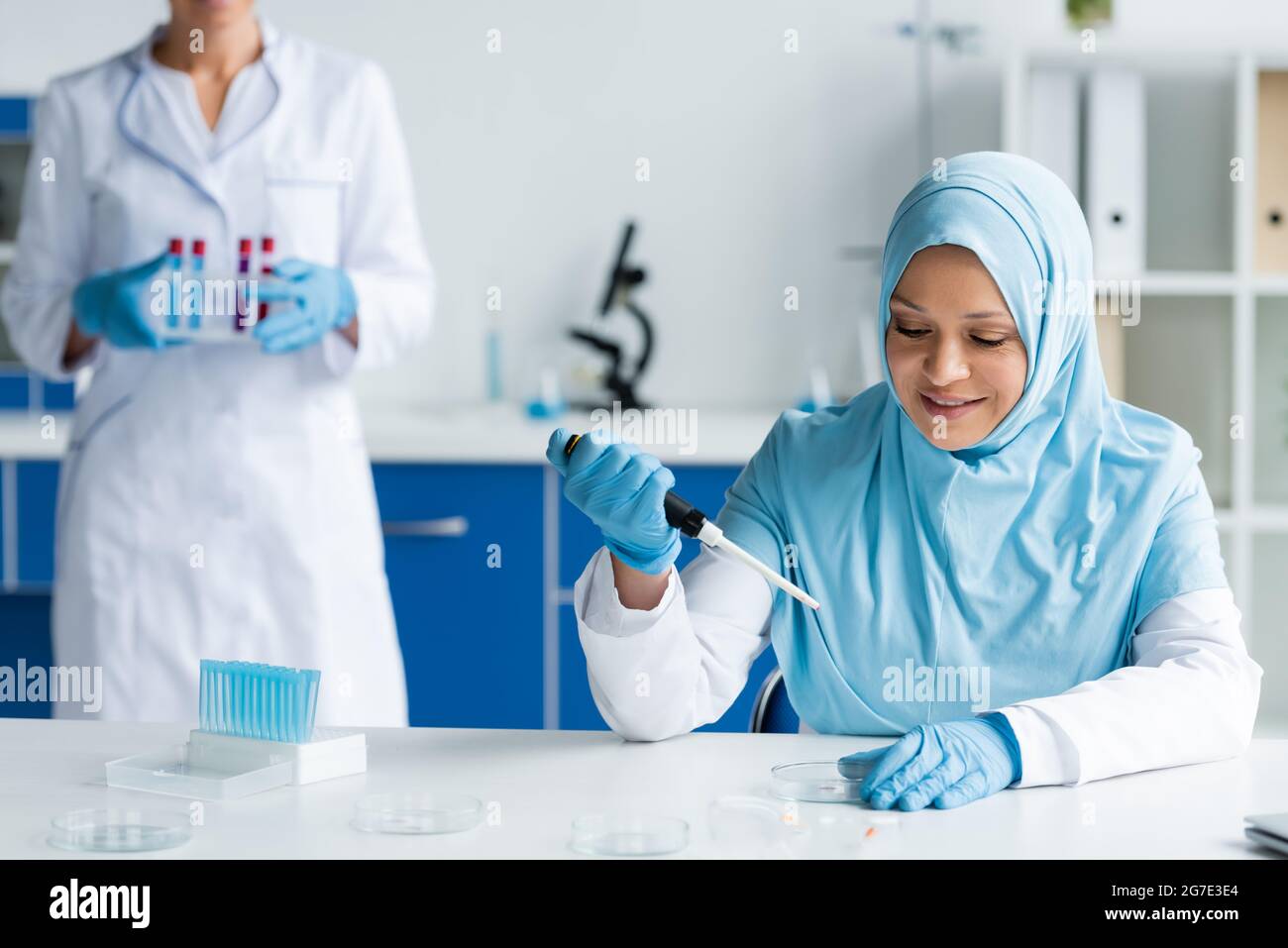 Sorridente scienziato arabo che tiene una pipetta elettronica vicino a piatti Petri e colleghi Foto Stock