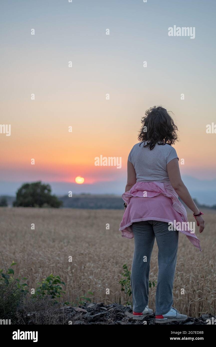 Immagine posteriore di una donna bruna in una T-shirt bianca a maniche corte e pantaloni grigi che guardano al tramonto e guardano l'orizzonte in un campo di cereali. Foto Stock