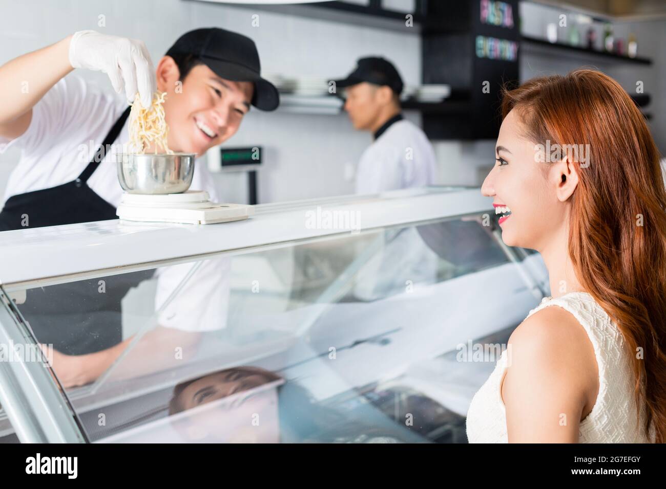 Ridendo giovane asiatico servendo una bella donna che pesa il formaggio a un banco di delicatessen con un sorriso amichevole Foto Stock