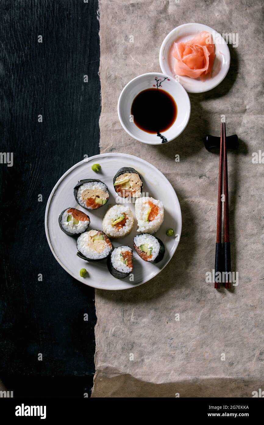 Involtini di sushi fatti in casa con salmone, omelette giapponesi, avacado, zenzero, wasabi e salsa di soia con chopsticks su carta grigia su sfondo di legno nero Foto Stock