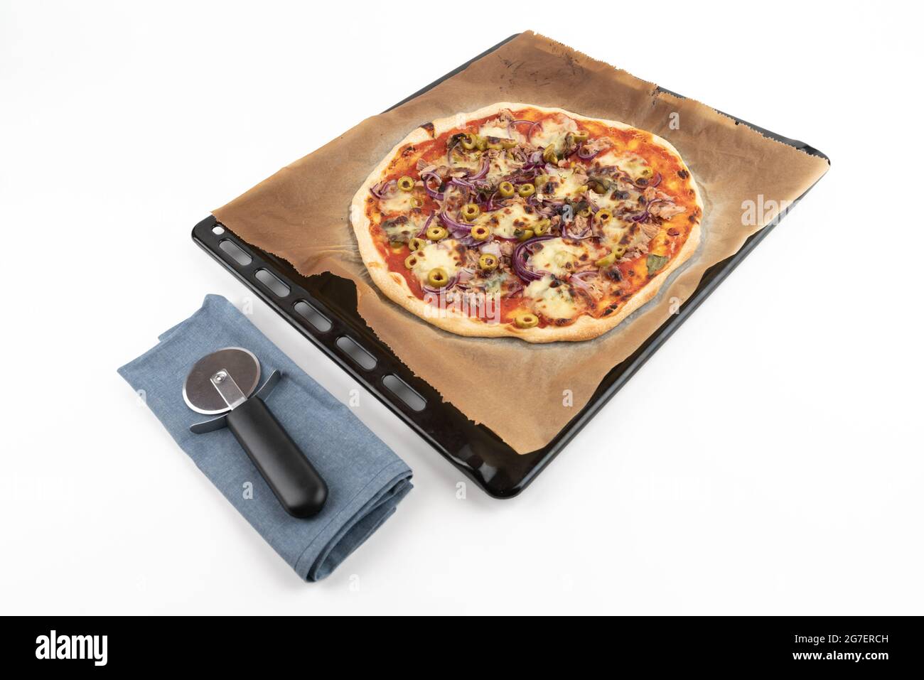 Foto in studio ad alto angolo di pizza fatta in casa al tonno su vassoio da forno nero e carta da forno, isolato su sfondo bianco. Panno blu e affettatrice per pizza Foto Stock