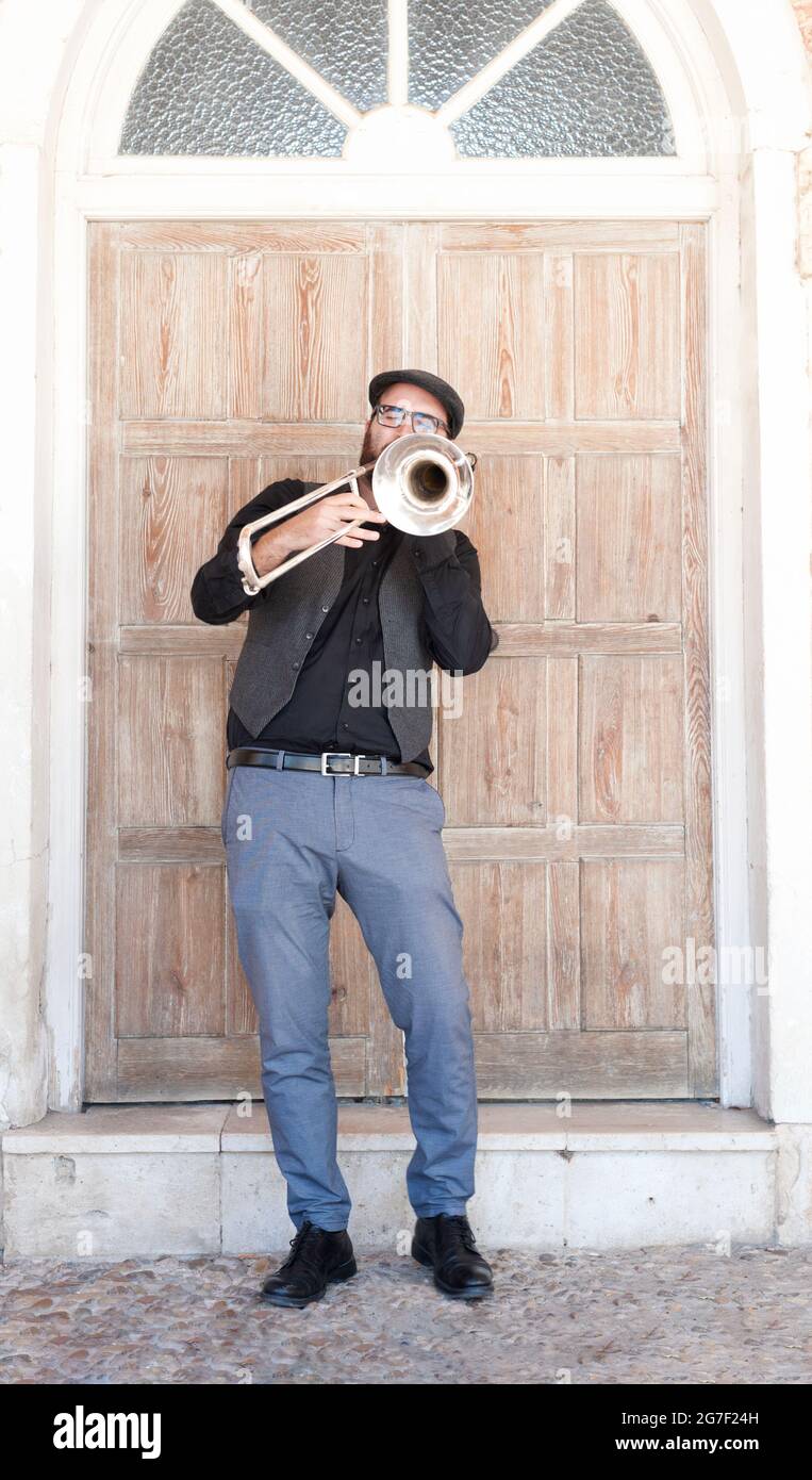 Musicista jazz con occhiali e barba che suona il trombone in piedi per strada. Vestito con un cappello inglese, una giacca, pantaloni blu. Dietro è un d di legno Foto Stock
