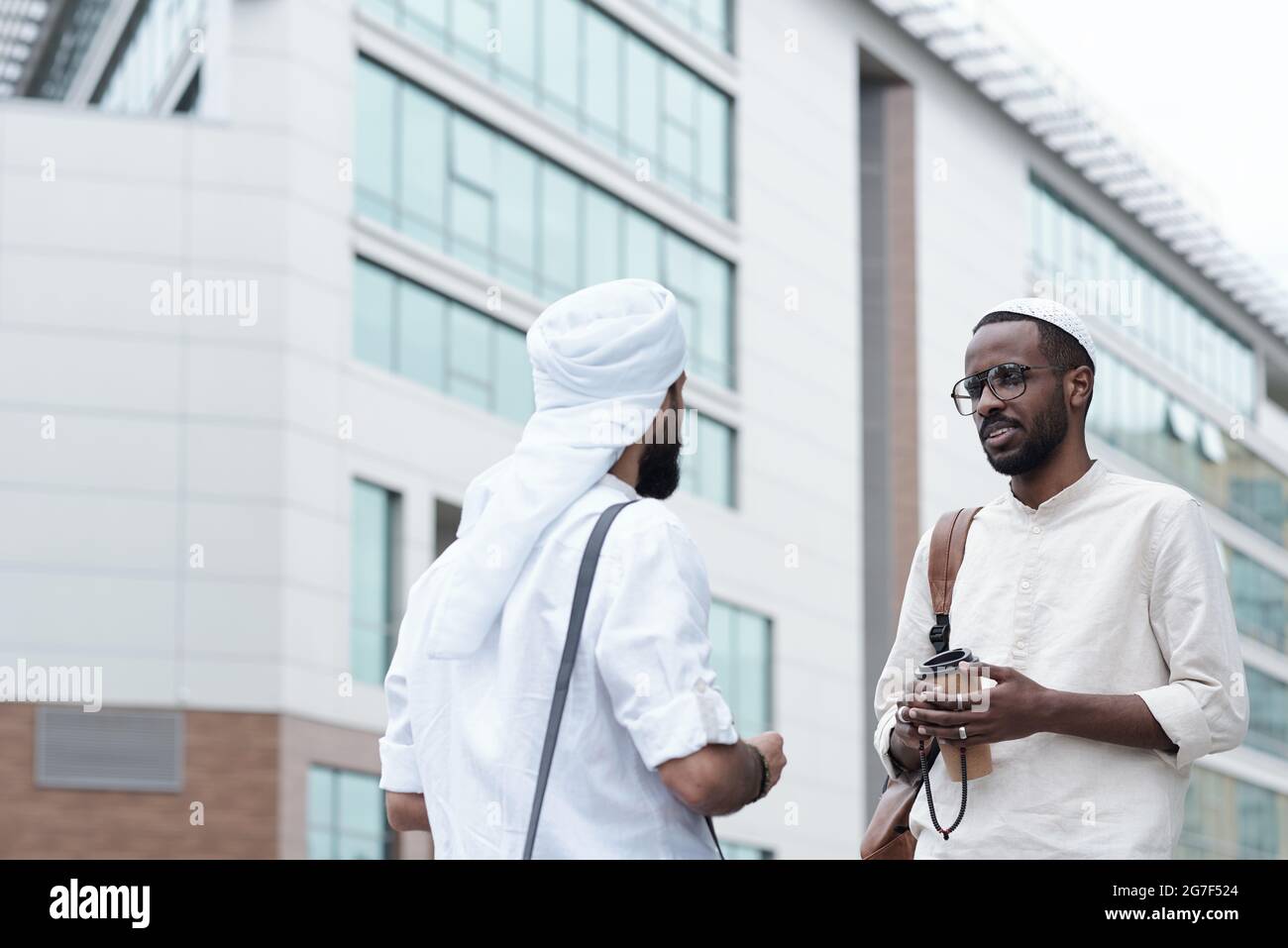 Soddisfare giovani uomini multietnici in abiti islamici in piedi contro l'edificio moderno e parlare di affari all'aperto Foto Stock