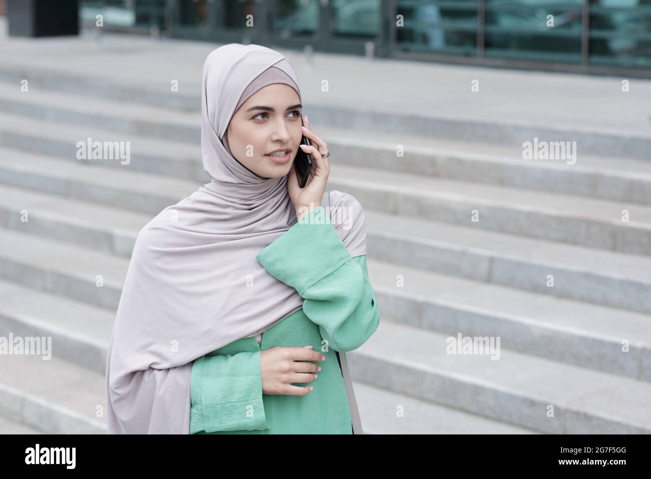 Giovane donna islamica fiduciosa in abito verde in piedi presso l'edificio dell'ufficio e che parla per telefono Foto Stock