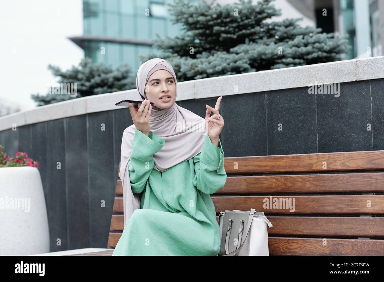 Giovane donna musulmana in abiti religiosi seduto sulla panchina e puntando con il dito mentre registra un messaggio audio sul telefono Foto Stock