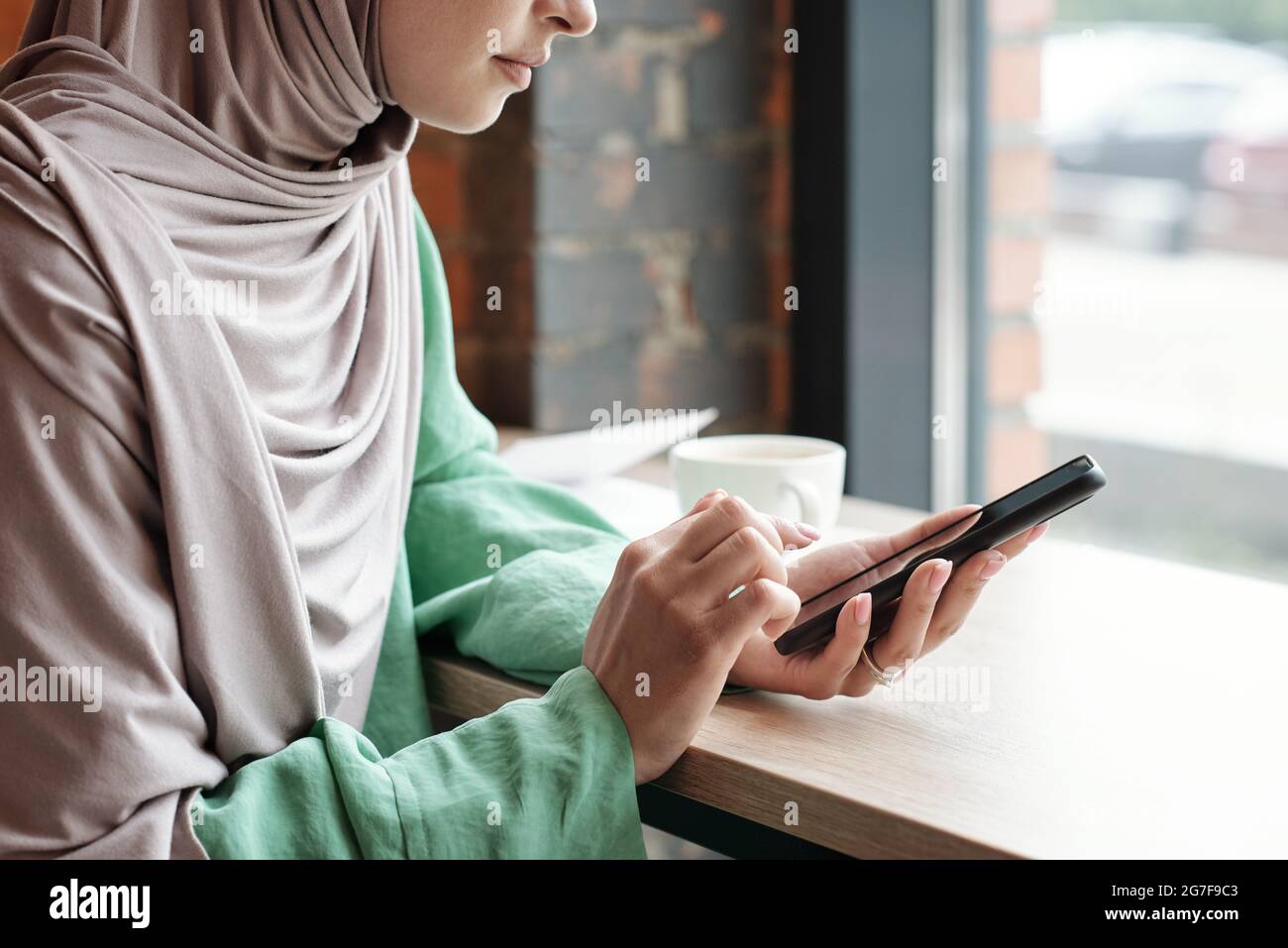Primo piano di calma giovane donna in hijab seduto al tavolo e rispondendo messaggi telefonici in caffetteria Foto Stock