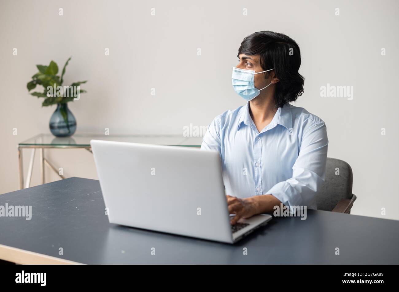 Giovane indiano che indossa una maschera medica protettiva utilizzando un computer portatile seduto alla scrivania in ufficio, il dipendente maschile guarda via con attenzione, preoccupandosi di crisi a causa della pandemia Foto Stock