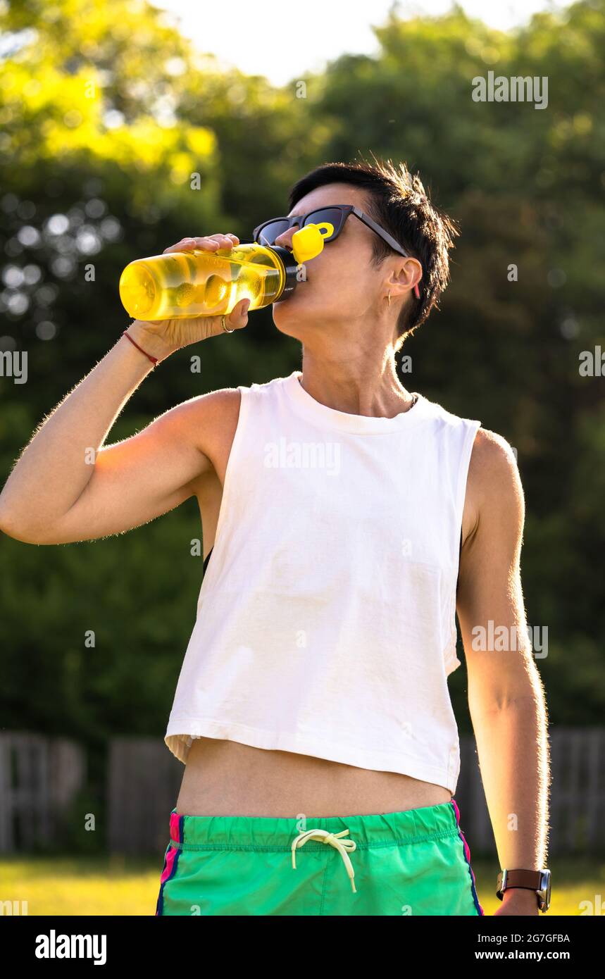 Giovane bella donna bere acqua dalla bottiglia di plastica in un parco dopo l'allenamento, capelli corti neri, occhiali da sole e pantaloncini, sport all'aperto in estate, Foto Stock