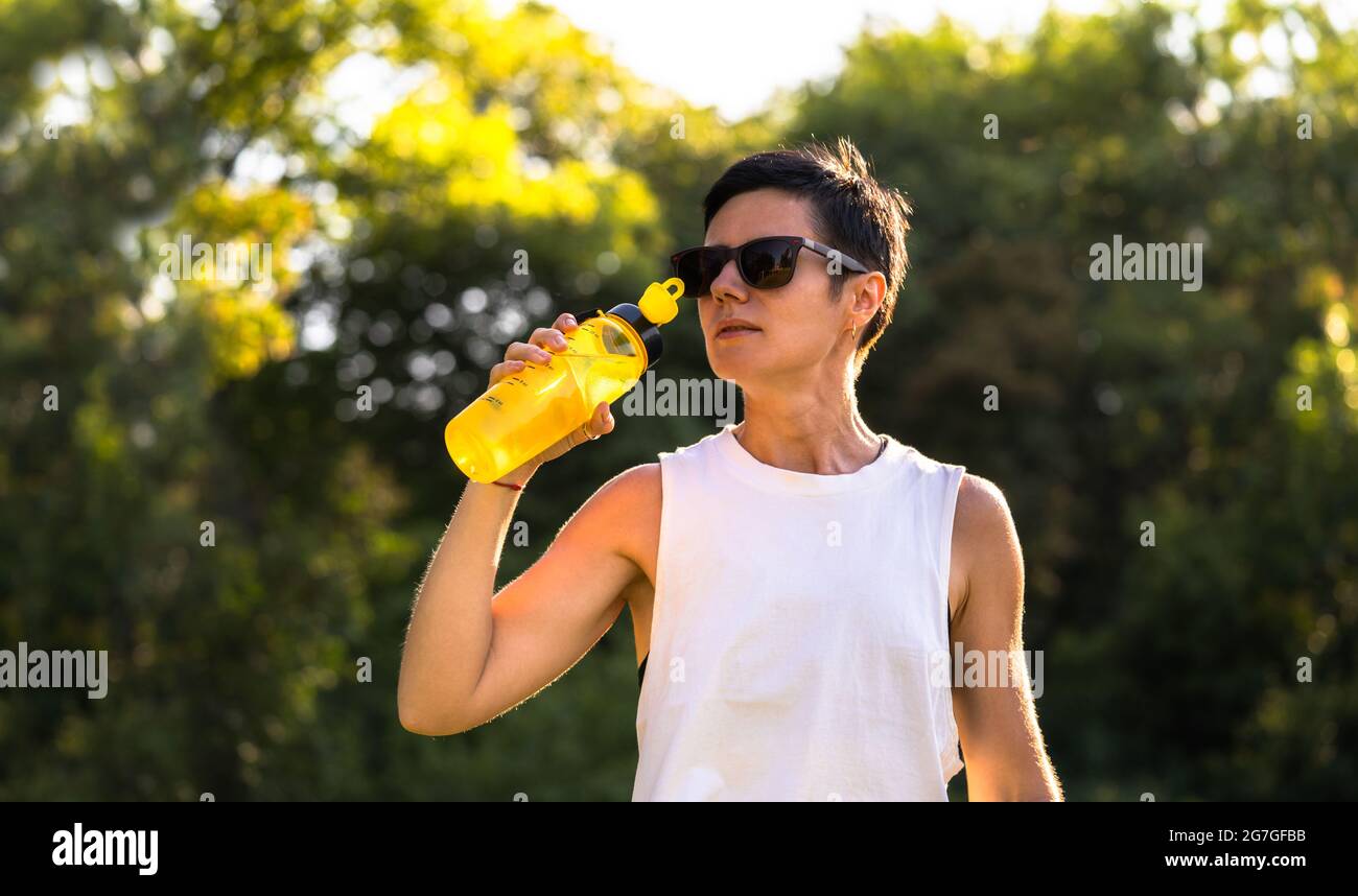 Giovane bella donna bere acqua dalla bottiglia di plastica in un parco dopo l'allenamento, capelli corti neri, occhiali da sole e pantaloncini, sport all'aperto in estate, Foto Stock