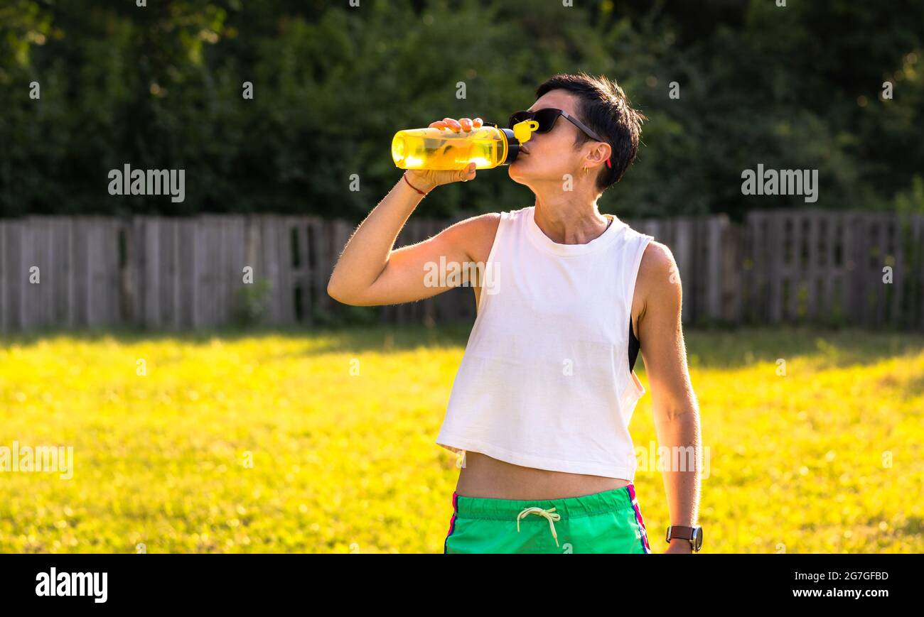 Giovane bella donna bere acqua dalla bottiglia di plastica in un parco dopo l'allenamento, capelli corti neri, occhiali da sole e pantaloncini, sport all'aperto in estate, Foto Stock