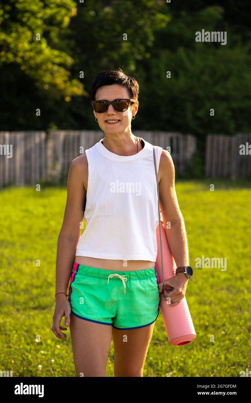Giovane bella donna con tappetino yoga in un parco, capelli neri corti,  occhiali da sole e pantaloncini, sport all'aperto in estate, ragazza  fitness sottile in abbigliamento sportivo, fem Foto stock - Alamy