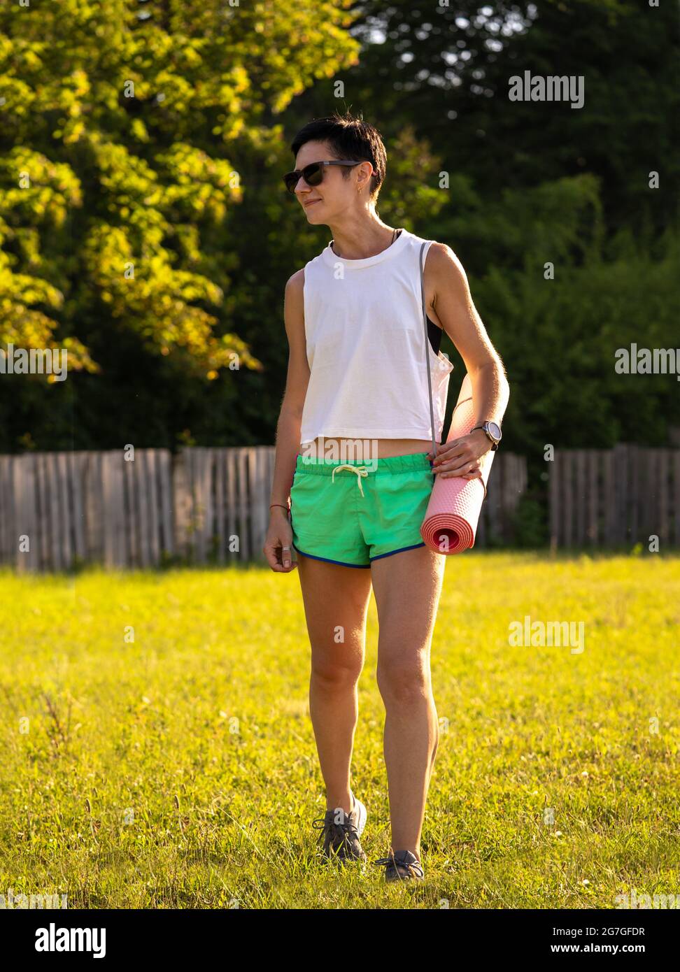 Giovane bella donna con tappetino yoga in un parco, capelli neri corti, occhiali da sole e pantaloncini, sport all'aperto in estate, ragazza fitness sottile in abbigliamento sportivo, fem Foto Stock