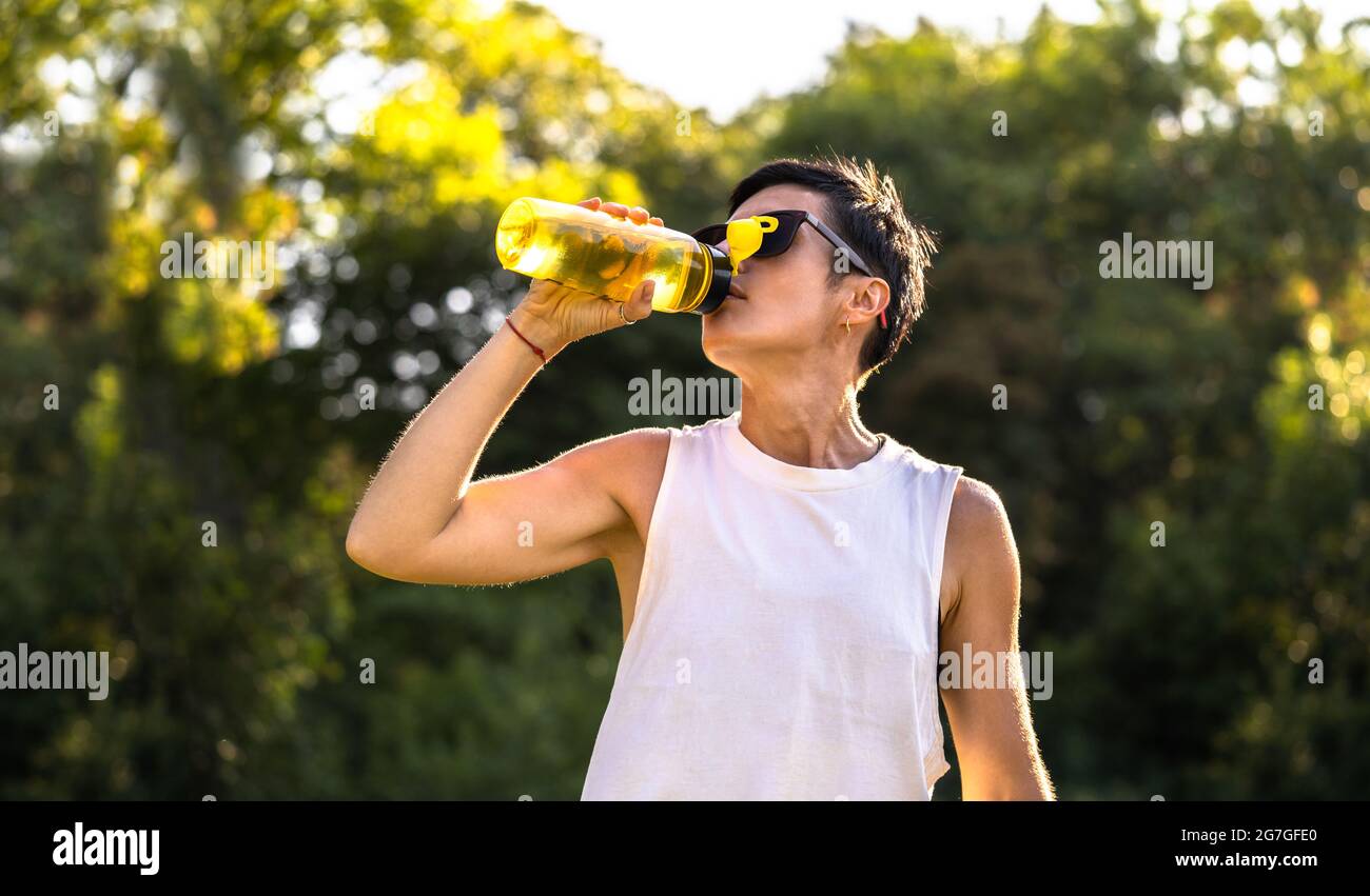 Giovane bella donna bere acqua dalla bottiglia di plastica in un parco dopo l'allenamento, capelli corti neri, occhiali da sole e pantaloncini, sport all'aperto in estate, Foto Stock