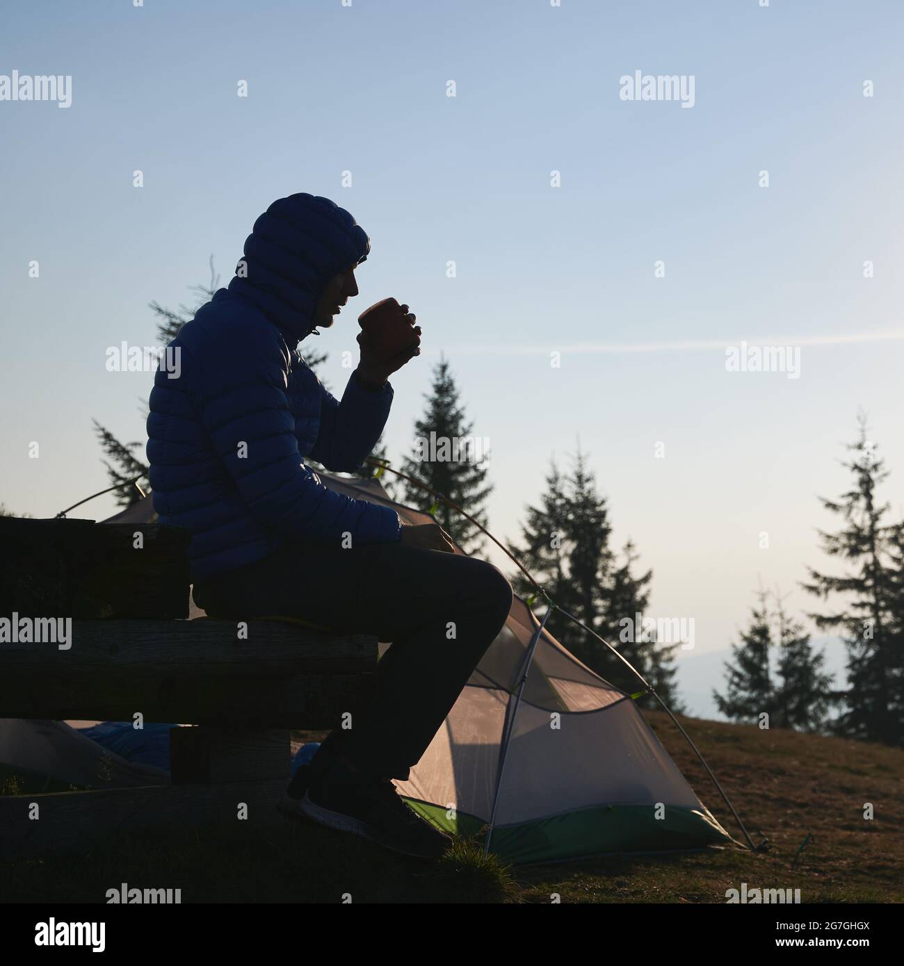 Silhouette di escursionista maschile riposato sulla panchina dopo una notte fresca in tenda, gustando un caffè profumato sullo sfondo di alberi solitari e cielo azzurro limpido e senza nuvole. Foto Stock