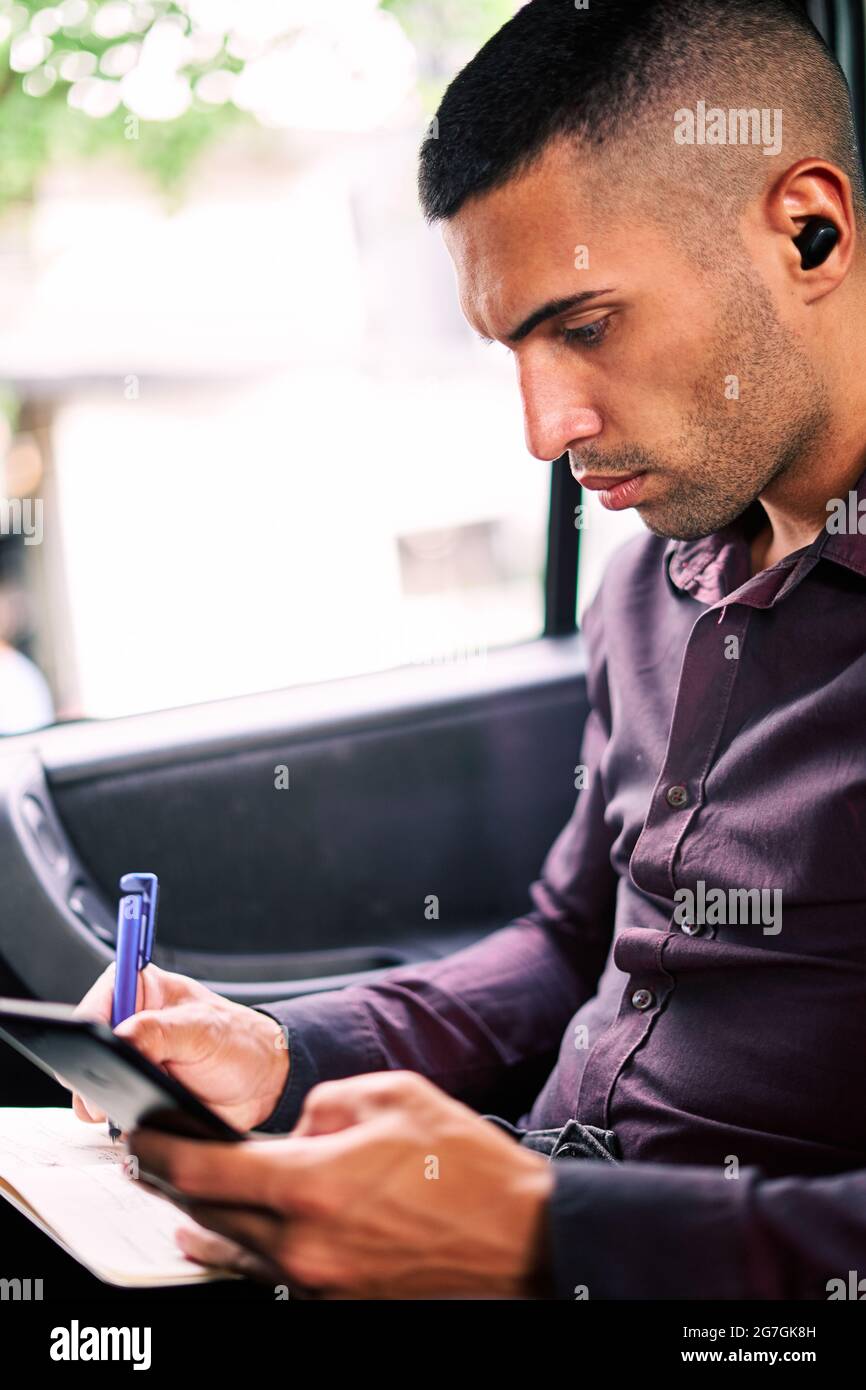 Imprenditore ispanico maschile messo a fuoco seduto sul sedile del passeggero dell'auto e scrivendo nel pianificatore mentre si è in viaggio di lavoro Foto Stock