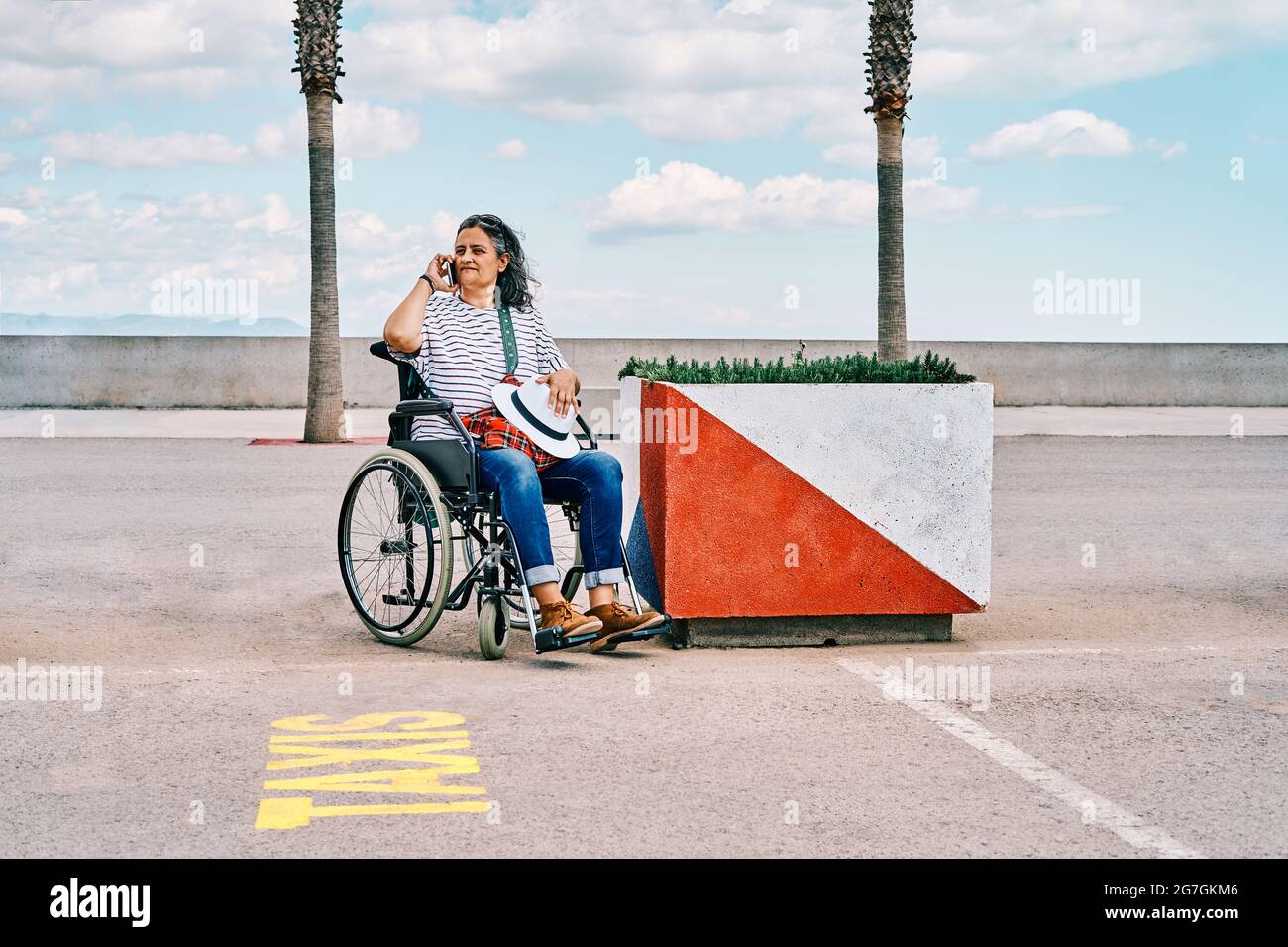 Donna mature disabili seduta in sedia a rotelle e che parla sul telefono cellulare mentre si chiama per il taxi sul parcheggio vicino argine Foto Stock