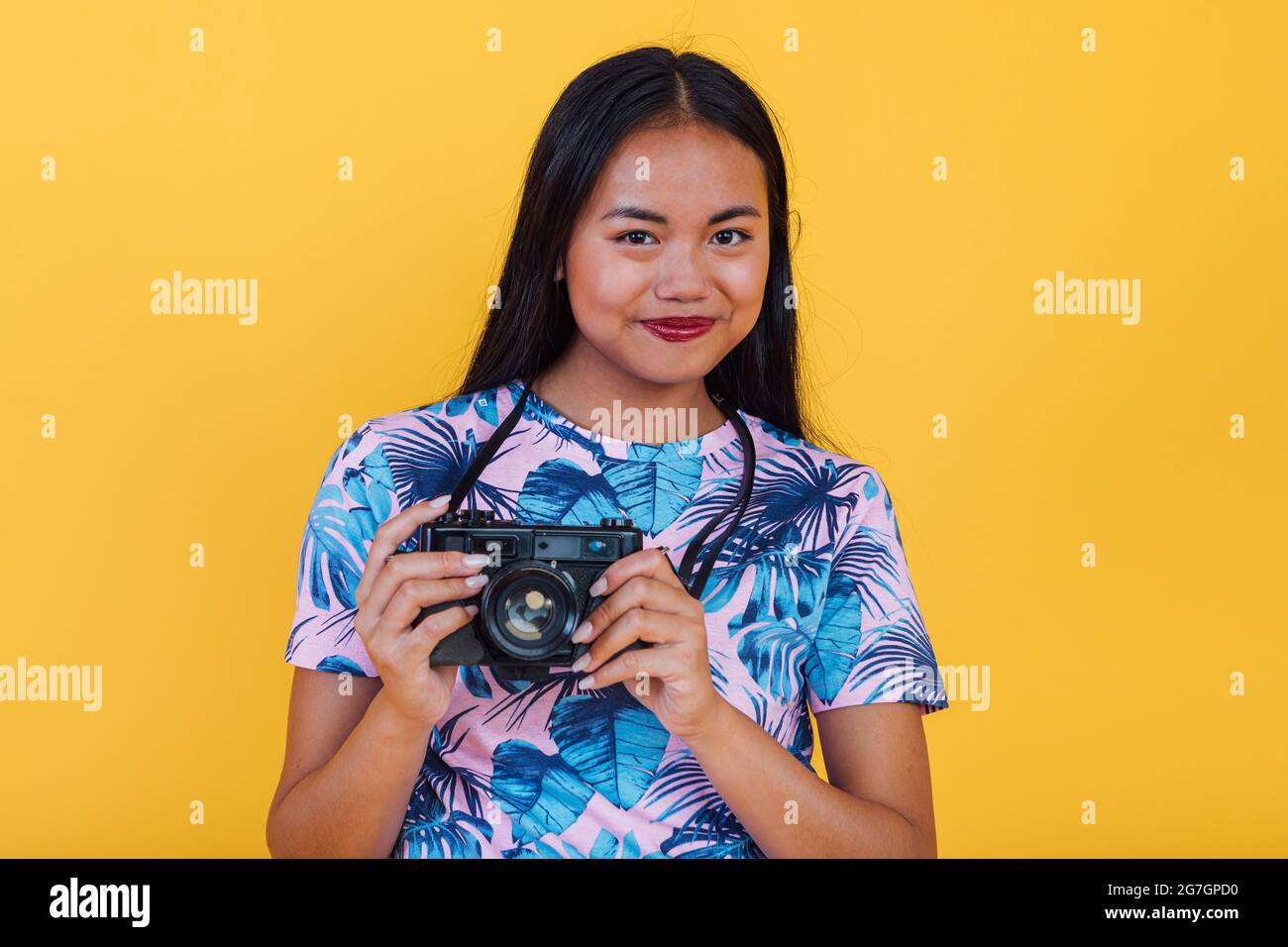 Donna asiatica felice in t shirt con stampa tropicale foglia che tiene fotocamera su sfondo giallo in studio Foto Stock